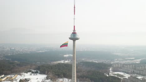 AERIAL:-Wide-Shot-of-Vilnius-Television-Tower-on-Sunny-Winter-Day-with-Waving-Lithuanian-Flag