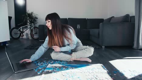 Young-woman-sitting-cross-legged-on-modern-apartment-floor-with-bicycle-on-background-playing-with-puzzle-and-chatting-on-laptop-device-in-leisure-time,-smart-lifestyle-caucasian-girl-slow-motion