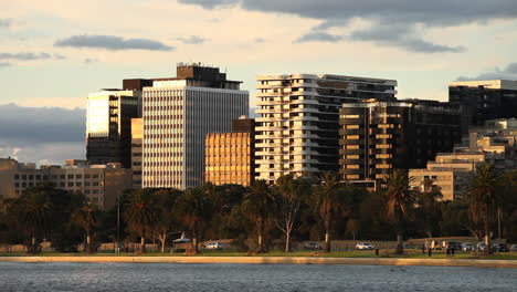 Modern-apartment-buildings-with-lake-view-reflective-golden-sunset-onto-traffic-and-recreational-walkers-below