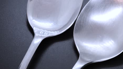 Close-up-view-of-two-steel-spoons-on-a-black-bacground,-rotating-motion