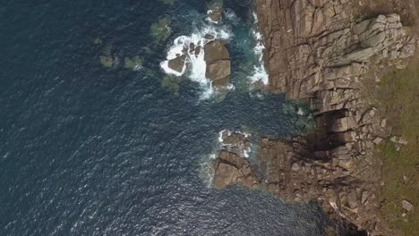 Vertical-aerial-over-rugged-stone-ocean-cliffs,-waves-crash-on-rocks