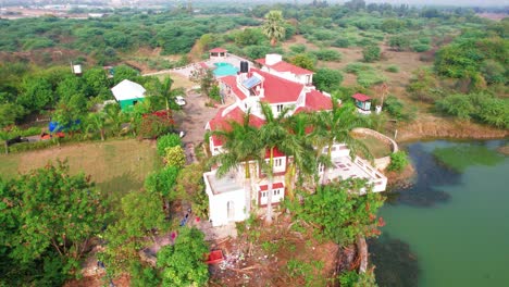 Soaring-up-drone-shot-of-a-vacation-resort-with-luxurious-white-houses,-lake-and-swimming-pool-in-Vadodara,-India,-surrounded-by-trees-and-green-vegetation