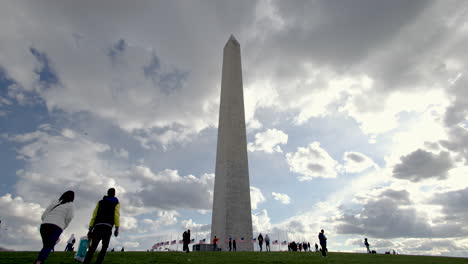 Group-of-people-visiting-the-Washington-Monument,-dramatic-cloudscape,-and-a-plane-in-distance---an-ultra-wide-shot