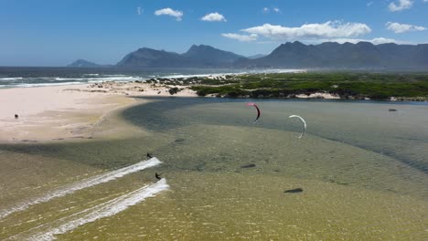 Wide-aerial-shot-of-a-pair-of-kite-surfers-racing-in-a-lagoon-near-a-sandy-beach-on-a-bright-sunny-day-with-beautiful-mountains-in-the-background-near-Hermanus-South-Africa