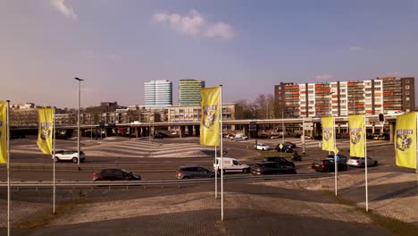 Aerial-with-banner-flags-of-local-Arnhem-city-Dutch-soccer-club-along-the-highway-coming-through-the-urban-hub-with-high-rise-residential-and-colorful-central-train-station-buildings-in-the-background