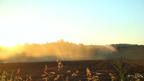 irrigation-and-Watering-Equipment-in-Farmland-Field,-Water-Splashing-and-Spraying-at-Sunset-on-Agricultural-Soil-in-Cingoli-Italy,-Panning-View