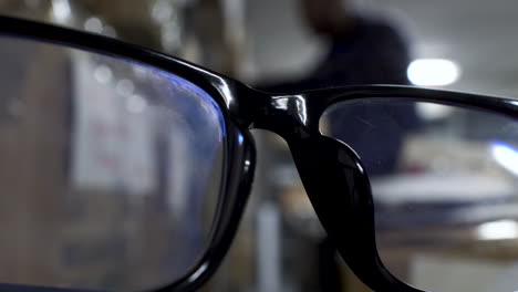 Nahaufnahme-Einer-Schwarzen-Randbrille-Mit-Bokeh-Hintergrund-Eines-Mannes,-Der-Durch-Regale-Sucht