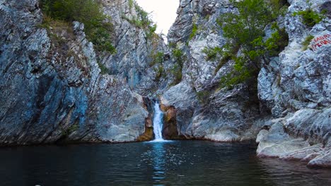 Wasserfall-Boaza-Wasserfall-Mit-Steinigen,-Mit-Bäumen-Bewachsenen-Klippen-Rechts-Mit-Einem-Kleinen-See-In-Zeitlupe-4k