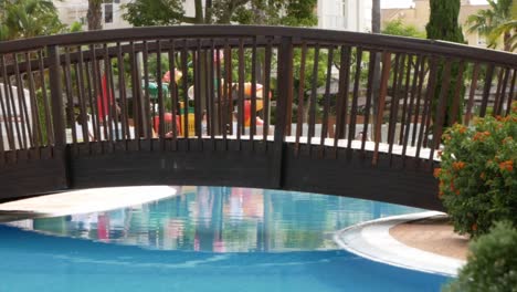 Peaceful-scene-of-Wooden-bridge-over-luxury-pool,-kids-Passing-in-Background