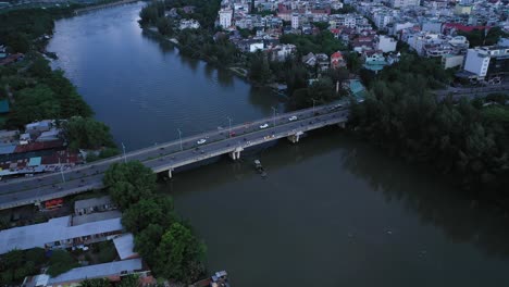Luftaufnahme-Eines-Flussbootes,-Das-Unter-Einer-Städtischen-Verkehrsbrücke-Und-Uferwohnungen-Entlang-Eines-Kanals-In-Ho-Chi-Minh-Stadt,-Vietnam,-Im-Nachmittagslicht-Hindurchfährt