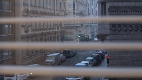 View-through-window-blind:-Snow-falls-on-narrow-old-world-city-street