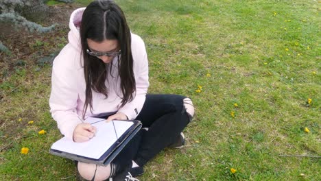 Junge-Teenager-Schriftstellerin-Mit-Brille-Schreibt-Mit-Einem-Stift-Auf-Ein-Blatt-Papier-Und-Sitzt-Draußen-Im-Gras