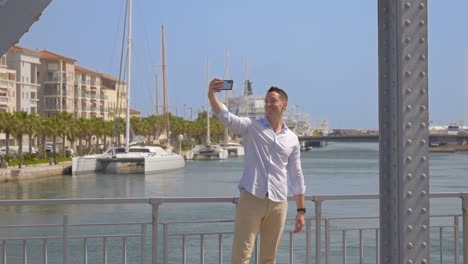 a-happy-young-man-takes-selfies-on-a-metal-bridge-with-passing-cars,-in-a-sunny-city-by-the-sea,-with-a-catamaran-in-the-background