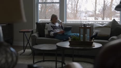 Woman-sitting-on-the-couch-in-front-of-a-bay-window,-drinking-coffee-while-scrolling-on-a-tablet-from-far-away