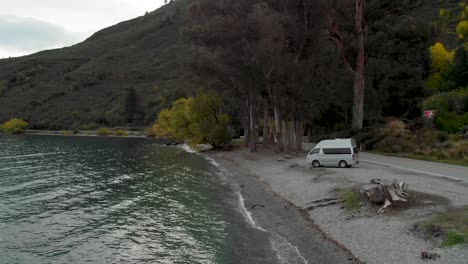 Motorhome-by-beautiful-blue-Lake-Wakatipu,-Queenstown,-New-Zealand-with-mountains-fresh-snow-cloudy-sky-in-background---Aerial-Drone