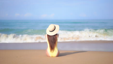 Back-of-Lonely-Woman-in-Swimsuit-Sitting-on-Sand-of-Tropical-Beach-in-Front-of-Sea-Waves-and-Blue-Horizon