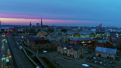 Luftdrohnenaufnahme-Von-Türmen-Entlang-Der-Skyline-Der-Stadt-Riga-Bei-Sonnenuntergang-Mit-Violettem-Und-Blauem-Himmel-über-Dem-Horizont