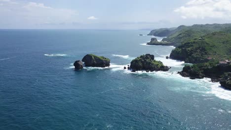 Drone-shot-of-overgrown-rocks-in-ocean,hit-by-waves-during-sunny-day---TIMANG-ISLAND,-YOGYAKARTA,-INDONESIA