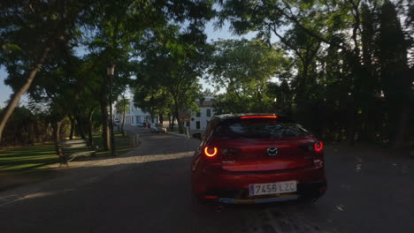 Dynamic:-FPV-drone-lines-up-behind-red-Mazda-car-driving-through-park