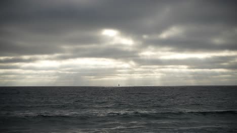 An-Einem-Düsteren-Tag,-An-Dem-Sonnenstrahlen-Durch-Die-Wolken-Scheinen,-Ist-Am-Horizont-Des-Ozeans-Das-Segel-Eines-Fernen-Segelboots-Zu-Sehen