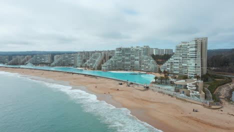 Aerial-dolly-out-of-sea-and-sand-shore-near-world-largest-swimming-pool-and-resorts-in-Algarrobo,-Chile