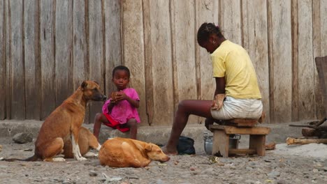 a-child-sits-next-to-three-dogs-and-eats-next-to-his-african-mother-on-the-ground-outside-while-she-stirs-the-pot---close-up