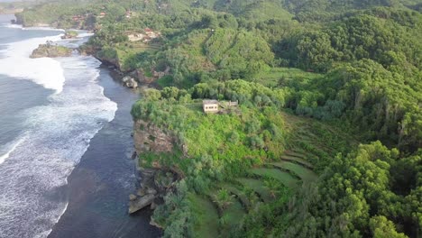 Luftaufnahme-Eines-Interessanten-Ortes-Mit-Wellen,-Die-Die-Grüne-Küste-Indonesiens-Erreichen,-Mit-Einem-Privathaus-Auf-Einer-Klippe