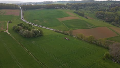 Weitwinkel-Drohnenaufnahmen-Eines-Traktors,-Der-Große-Grüne-Weiden-Neben-Einer-Asphaltstraße-Besprüht
