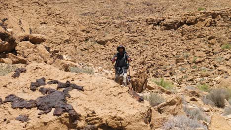 One-man-trekker-with-trekking-poles-walking-down-the-sandstone-cliffy-slope-during-travel-in-Ramon-Crater-Trail,-Negev-desert,-Israel