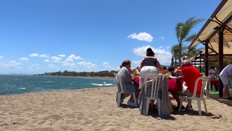 Family-sitting-at-the-beach-and-having-a-meal-in-a-restaurant-with-a-sea-view-on-a-windy-day-in-Marbella-Spain,-sunny-weather-and-blue-sky-with-palm-trees,-4K-shot