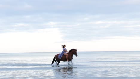 A-beautiful-girl-with-long-hair-in-a-blue-dress-riding-a-horse-through-the-water-during-the-evening,-Donabate,-Ireland