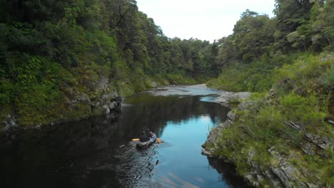 SLOWMO---Two-males-paddle-canoe-through-rocky-canyon-on-Pelorus-river,-New-Zealand-with-rock-boulders-and-forrest-in-background---Aerial-Drone
