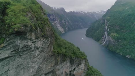 Aerial-Slomo-of-Geiranger-Fjord,-Norway,-flying-forwards-with-a-Moving-Boat-in-the-Background,-passing-The-Seven-Sisters