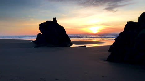 Two-people-sitting-on-a-tall-rock-at-the-beach-in-Bandon-Oregon,-enjoying-the-sunset