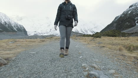 A-low-to-high-angle-follow-shot-of-a-woman-hiking-between-snow-capped-mountains-in-New-Zealand-on-a-cold-winters-morning