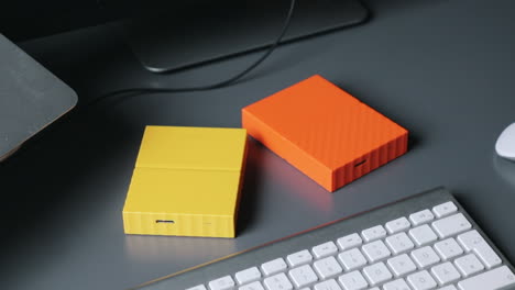 Male-hands-setting-two-yellow-and-orange-coloured-hard-drives-on-a-grey-table-midst-keyboard-and-mouse