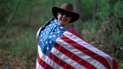 Woman-in-cowboy-hat-and-sunglasses-holding-out-an-American-flag-and-holding-it-up-to-her-body-and-laughing