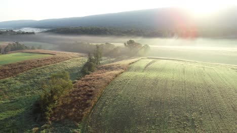Aerial-camera-moving-to-the-right-showing-farm-fields,-trees,-mountains,-and-fog-in-the-morning-light