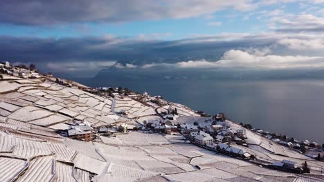 Vista-Aérea-Sobre-Un-Típico-Pueblo-Suizo-En-Lavaux-Viñedo-Cubierto-De-Nieve,-Los-Alpes-Y-El-Lago-Leman-En-El-Fondo