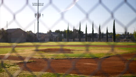 Slide-up-shot-looking-at-an-empty-green-baseball-field-diamond-from-behind-home-plate-and-a-chain-link-fence-in-a-public-park-in-the-early-morning
