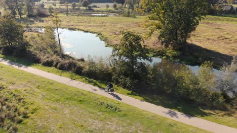 Ein-Einzelner-Radfahrer-Fährt-Durch-Eine-Landschaft-Mit-Grasfeldern-Und-Bäumen-Entlang-Der-Straße,-Daneben-Schlängelt-Sich-Ein-Fluss