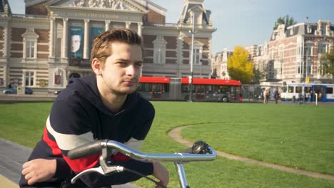 Handsome-young-man-upholding-on-a-bicycle-standing-near-the-Concert-Hall-in-Amsterdam-on-a-sunny-day