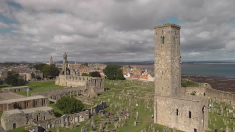 Aerial-drone-flying-by-one-of-the-towers-of-the-St-Andrews-Cathedral-as-other-towers-and-the-coastline-come-into-view-in-the-background-in-St-Andrews,-Scotland,-UK