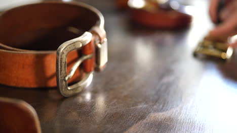 Close-up-on-a-leather-workers-workbench-with-worn-leather-belts-with-heavy-patina-on-the-buckle-as-he-works-on-a-new-belt