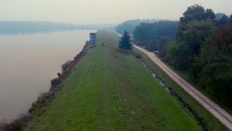 Aerial-view-of-a-man-standing-next-to-a-metal-silo-on-a-grassy-hill-overlooking-a-foggy-reflective-lake
