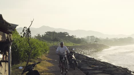 A-man-rides-a-cycle-near-a-beach-or-coast-of-an-ocean-at-sunset-or-sunrise-in-Pantai-Lebih,-Indonesia