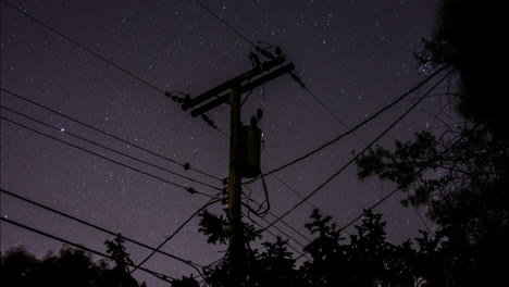 Ein-Zeitraffer-Der-Am-Himmel-Rotierenden-Sterne-Hinter-Einem-Silhouettierten-Telefonmast-In-Einem-Vorort-Sowie-Stromleitungen-Und-Bäumen-Am-Nachthimmel