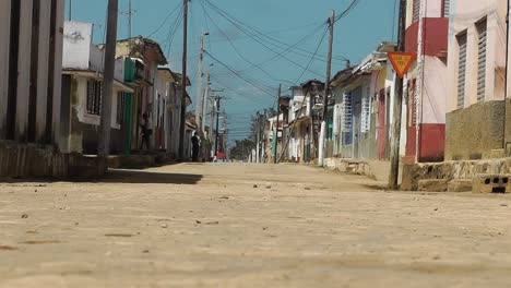 Low-angle-view-of-a-dirt-street-in-a-typical-Cuban-town-where-people-and-a-truck-are-passing-by-the-traditional,-colorful-houses-on-a-hot-and-sunny-day