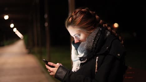 Mujer-Joven-Mirando-Su-Teléfono-Inteligente-Por-La-Noche-En-Un-Frío-Banco-Del-Parque
