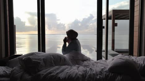 Woman-sips-coffee-and-looks-out-at-the-sunrise-from-her-bedroom-at-luxury-villa